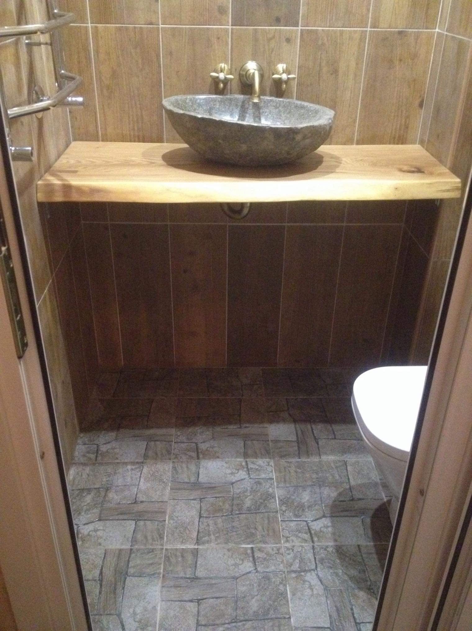 Łazienka z kamienną umywalką na blacie z surowego drewna, brązowe płytki na ścianach i podłodze imitujące kamień, widok zza uchylonych drzwi.