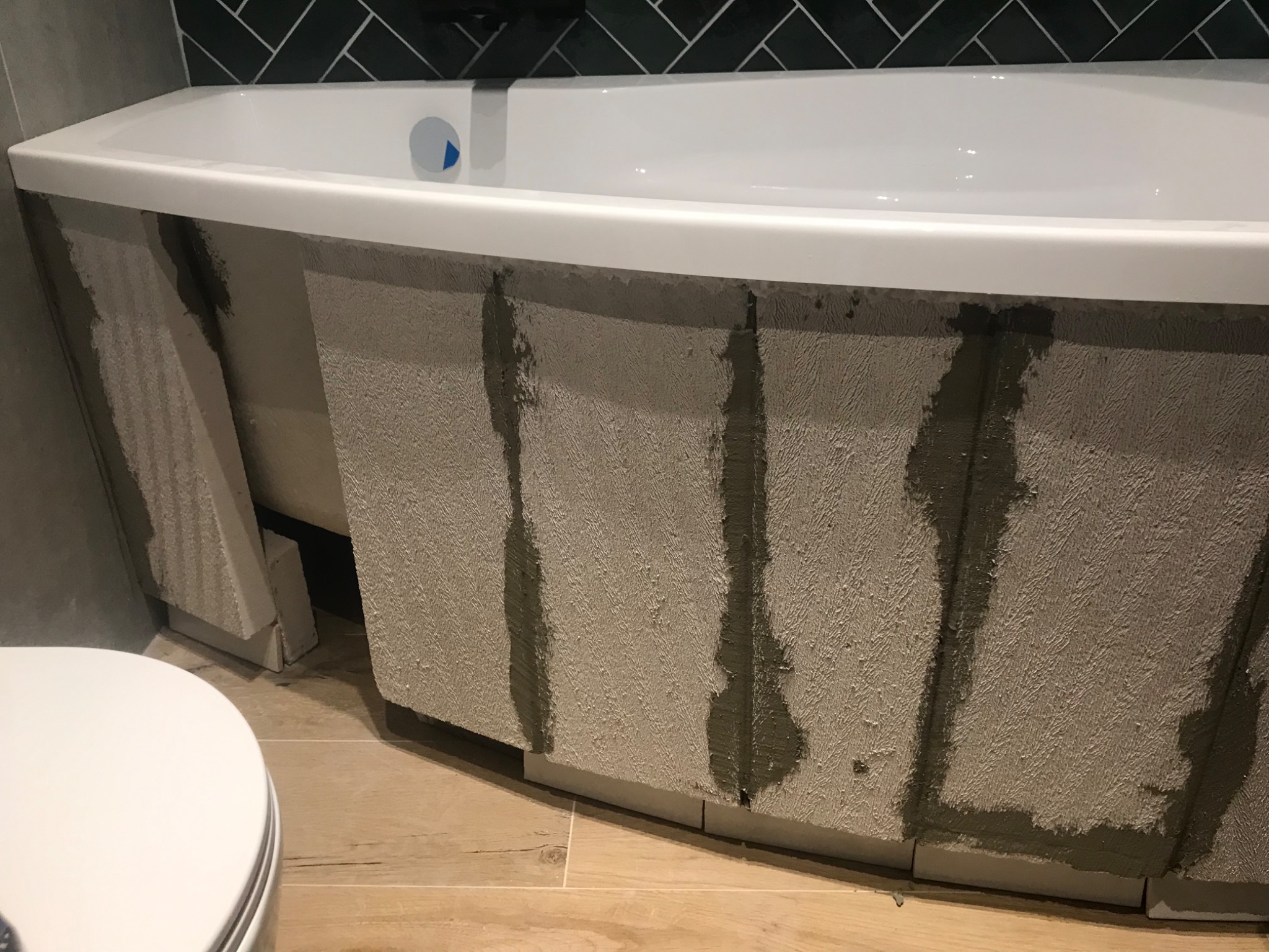 Wanna z obudową w trakcie wykańczania, widoczne fragmenty płytek i zaprawy klejowej, na podłodze panele imitujące drewno, w tle czarne płytki w jodełkę.