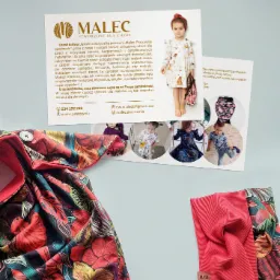 Kompozycja produktowa: ulotka reklamowa pracowni krawieckiej Malec z wizerunkiem dziecka w sukience i przykładami ubrań, obok leżą fragmenty kolorowych tkanin z motywem kwiatowym i czerwona opaska.