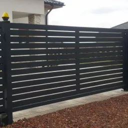 Nowoczesna, przesuwna brama wjazdowa z poziomych profili w kolorze antracytowym, z żółtą lampą ostrzegawczą na słupku, na tle domu z jasną elewacją.