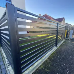 Nowoczesne, poziome przęsła ogrodzeniowe w kolorze grafitowym, oddzielające posesję od drogi, z widocznym trawnikiem i fragmentem domu w tle.
