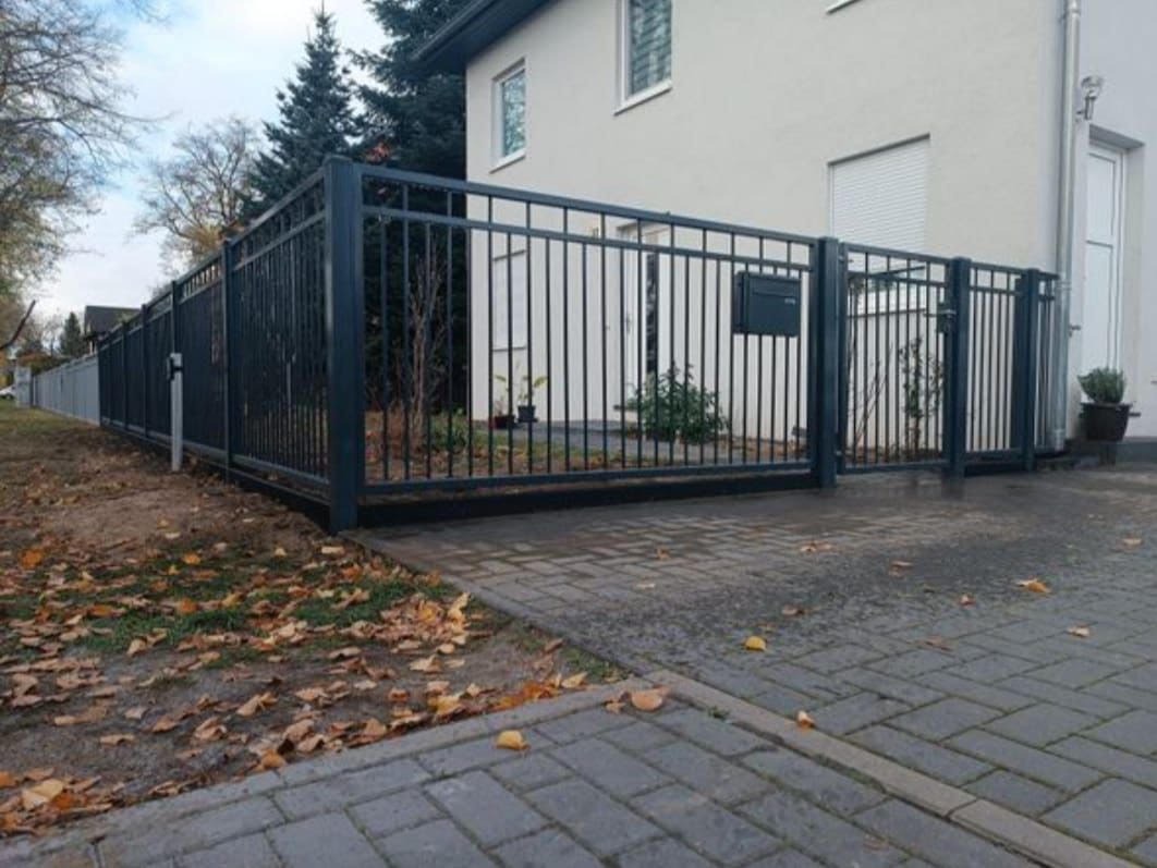 Nowoczesne, metalowe ogrodzenie posesji z przesuwną bramą wjazdową, widok z zewnątrz na tle domu z szarą kostką brukową i opadłymi liśćmi.