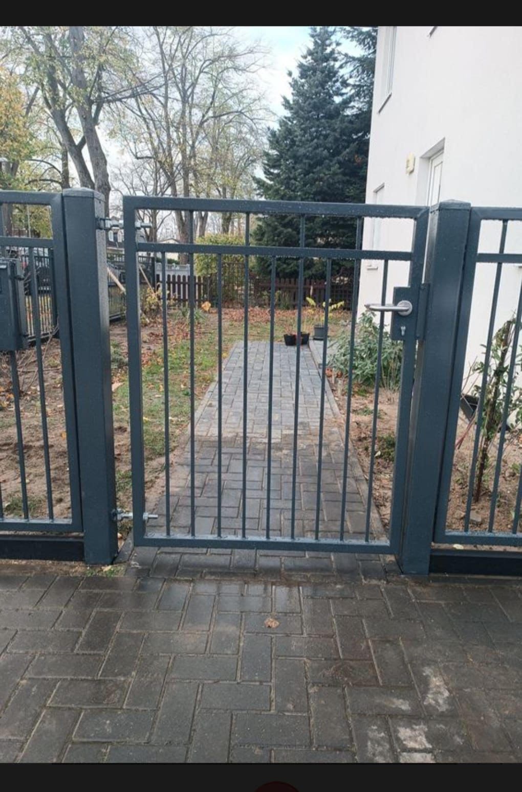 Metalowa furtka wejściowa w kolorze grafitowym, widok z zewnątrz na ścieżkę z kostki brukowej prowadzącą do domu.