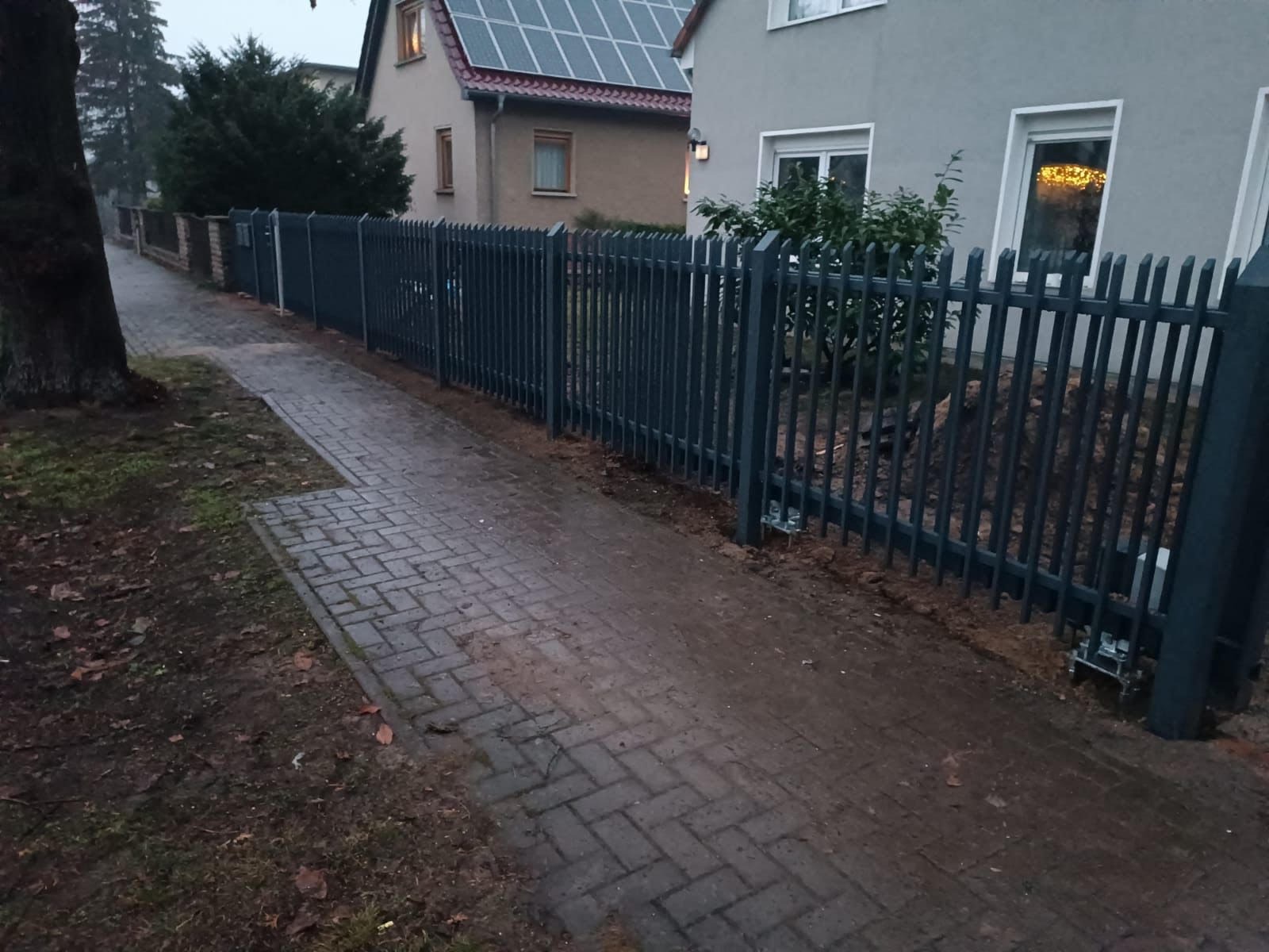 Nowoczesne ogrodzenie metalowe z pionowych prętów, pomalowane na ciemny szary kolor, zamontowane wzdłuż ścieżki z kostki brukowej, widoczne elementy mocujące do podłoża.