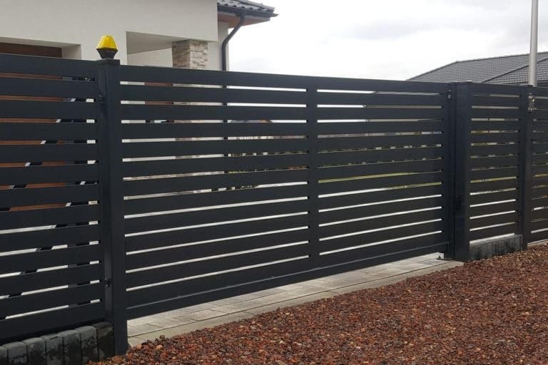 Nowoczesna, przesuwna brama wjazdowa z poziomych profili w kolorze antracytowym, z żółtą lampą ostrzegawczą na słupku, na tle domu z jasną elewacją.