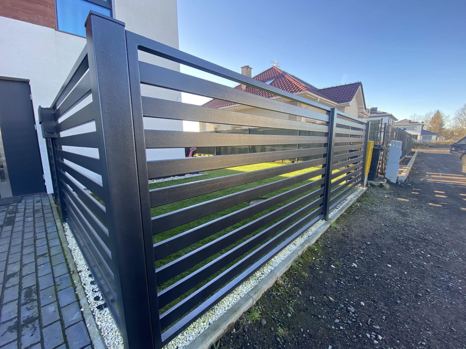 Nowoczesne, poziome przęsła ogrodzeniowe w kolorze grafitowym, oddzielające posesję od drogi, z widocznym trawnikiem i fragmentem domu w tle.