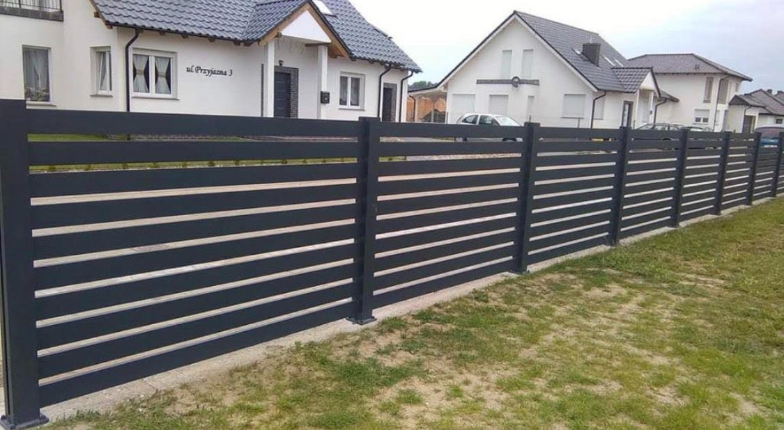 Nowoczesne ogrodzenie metalowe w kolorze grafitowym z poziomymi szczeblami, na tle białych domów jednorodzinnych z szarymi dachami.