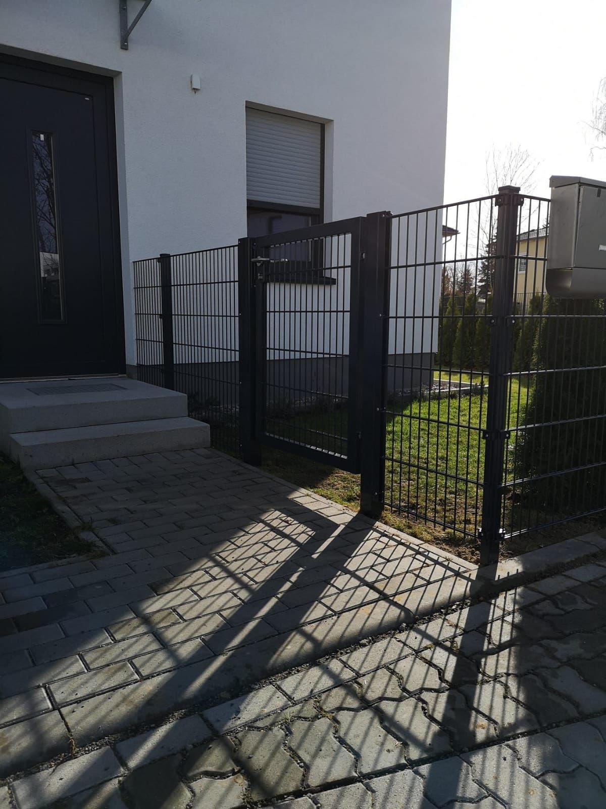 Nowoczesne, czarne ogrodzenie panelowe z furtką przy wejściu do domu z szarą kostką brukową i widocznymi cieniami