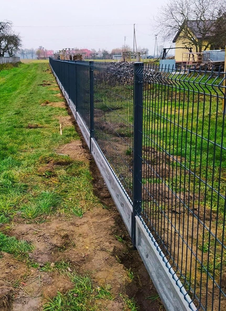 Długa, ciemna, panelowa konstrukcja z drutu na betonowej podmurówce, oddzielająca trawnik od pola, z domem w tle.