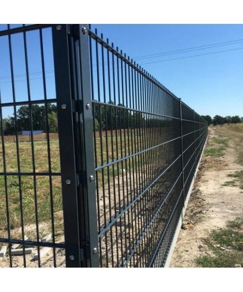 Długi, ciemny, panelowy system ogradzający z metalu, zamontowany na betonowej podstawie, widziany z boku na tle pola i błękitnego nieba.