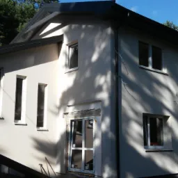 Dwukondygnacyjny budynek w trakcie wykończenia elewacji zewnętrznej, widoczne okna z białymi ramami i niezakończony montaż stolarki okiennej przy wejściu, cień drzew na ścianie.