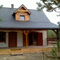 Ukończony dom z drewnianą werandą i ciemnym dachem na tle zieleni. Widoczne drewniane okiennice i elementy wykończeniowe. Styl rustykalny, naturalne materiały.