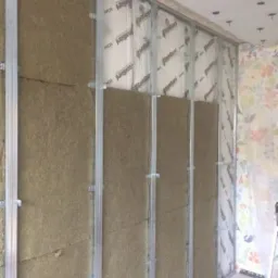 Montaż izolacji z wełny mineralnej w metalowym stelażu na ścianie z fragmentami tapety w kwiaty, widoczne białe membrany paroizolacyjne.