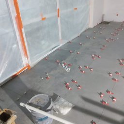 M-BUD - Szara podłoga w trakcie układania płytek, widoczne czerwone klipsy poziomujące, wiadro z zaprawą i worek z krzyżykami dystansowymi, ściany zabezpieczone folią malarską.
