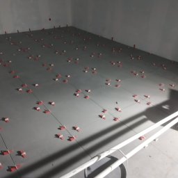M-BUD - Szare płytki podłogowe w trakcie układania, z czerwonymi klipsami poziomującymi, widoczne rurki PCV i wiadro z klejem w rogu pomieszczenia.