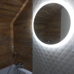 M-BUD - Nowoczesna łazienka z drewnopodobnymi płytkami na skośnej ścianie, białą wanną i okrągłym lustrem z podświetleniem LED na ścianie z geometrycznymi płytkami.