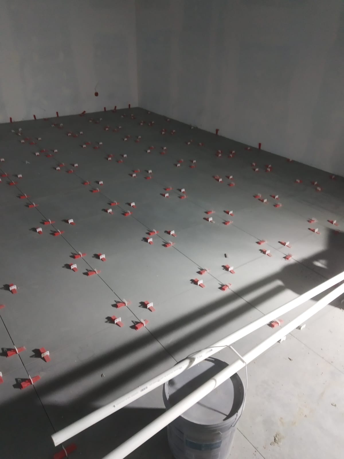 Szare płytki podłogowe w trakcie układania, z czerwonymi klipsami poziomującymi, widoczne rurki PCV i wiadro z klejem w rogu pomieszczenia.