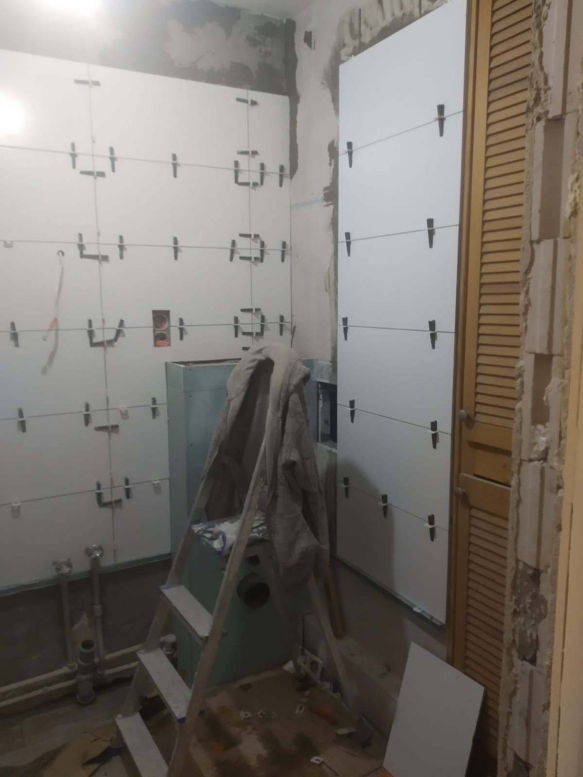 Białe płytki układane na ścianie łazienki, z widocznymi krzyżykami dystansowymi, drabina oparta o ścianę, fragment drewnianych drzwi po prawej stronie.