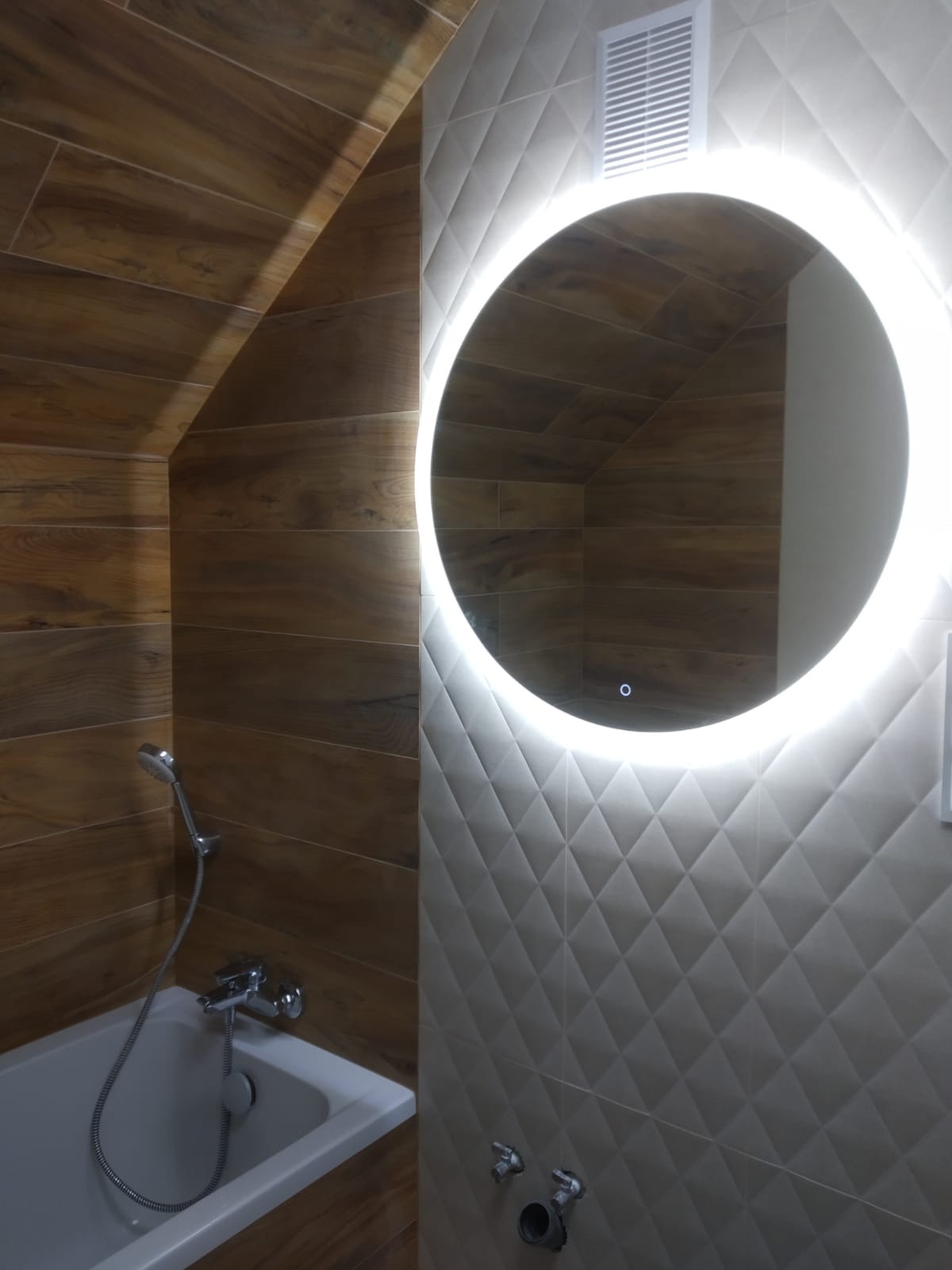 Nowoczesna łazienka z drewnopodobnymi płytkami na skośnej ścianie, białą wanną i okrągłym lustrem z podświetleniem LED na ścianie z geometrycznymi płytkami.