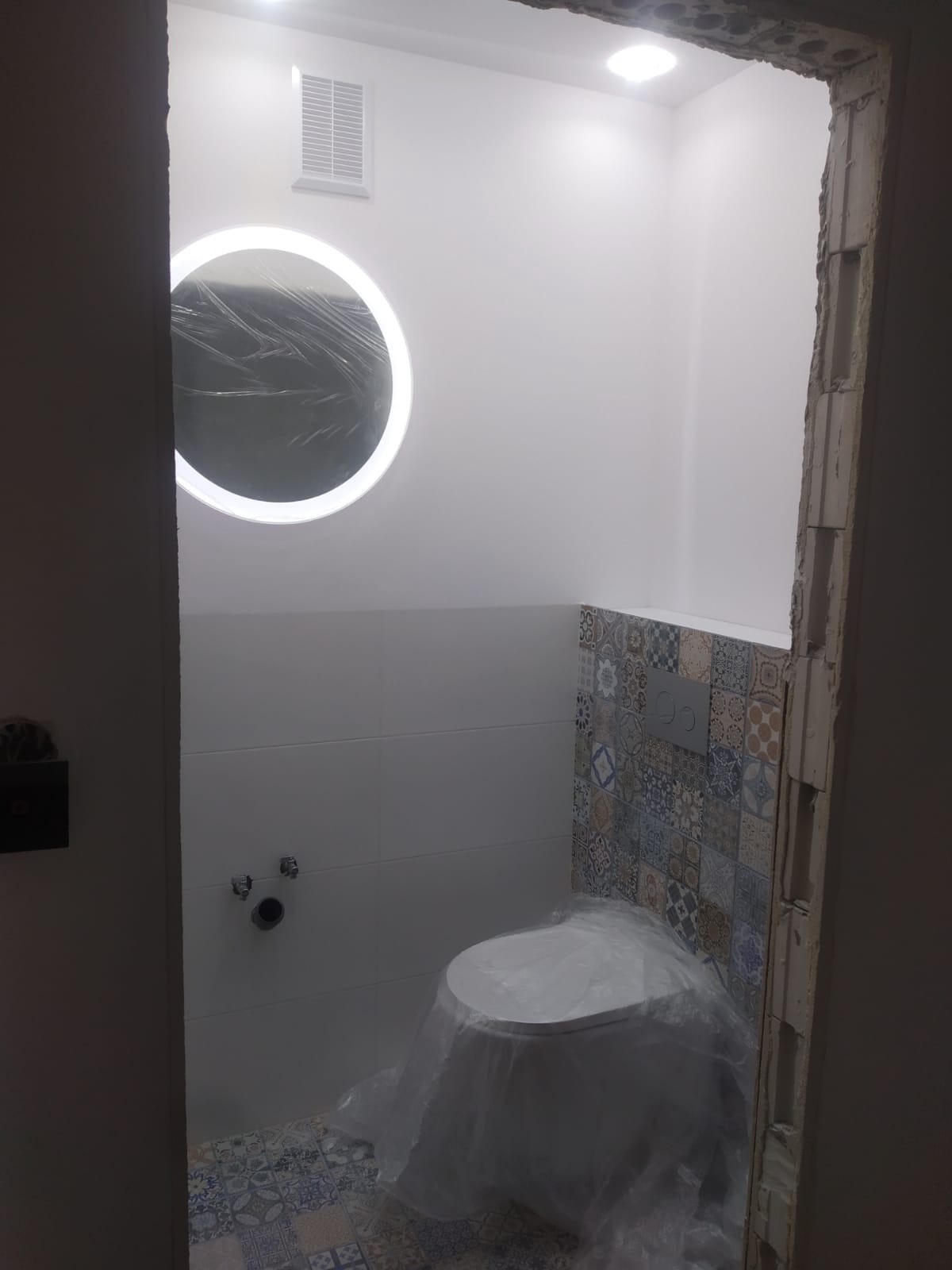 Nowoczesna łazienka w trakcie wykończenia, widoczna wisząca toaleta zabezpieczona folią, podświetlane okrągłe lustro, białe płytki ścienne i dekoracyjne płytki patchworkowe na ścianie nad toaletą...