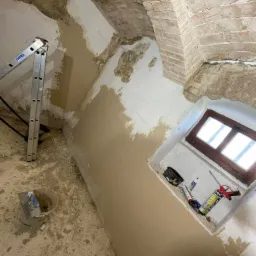 Wnętrze w trakcie prac: częściowo nałożona warstwa szpachli na ścianie z fragmentami cegieł, drabina Krause oparta o ścianę, wiadro z narzędziami na podłodze, tuby z silikonem przy oknie.