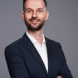 Portret uśmiechniętego mężczyzny z brodą w ciemnym garniturze i białej koszuli na szarym tle, pozującego z założonymi rękami.