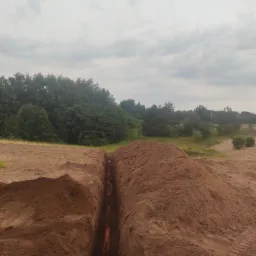 Długi, wąski wykop w ziemi, z widocznymi pomarańczowymi rurami na dnie, otoczony hałdami ziemi, z drzewami i pochmurnym niebem w tle.