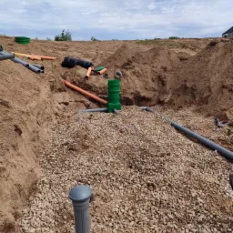 Fragment instalacji przydomowej oczyszczalni ściek&oacute;w w trakcie montażu: rury rozsączające, studzienka rozdzielcza, warstwa żwiru drenażowego w wykopie, widoczne elementy systemu rozsączania...