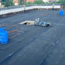 Czarny dach pokryty papą termozgrzewalną w trakcie prac dekarskich, widoczne butle gazowe, rolki papy, puszka z klejem i otwór w dachu.