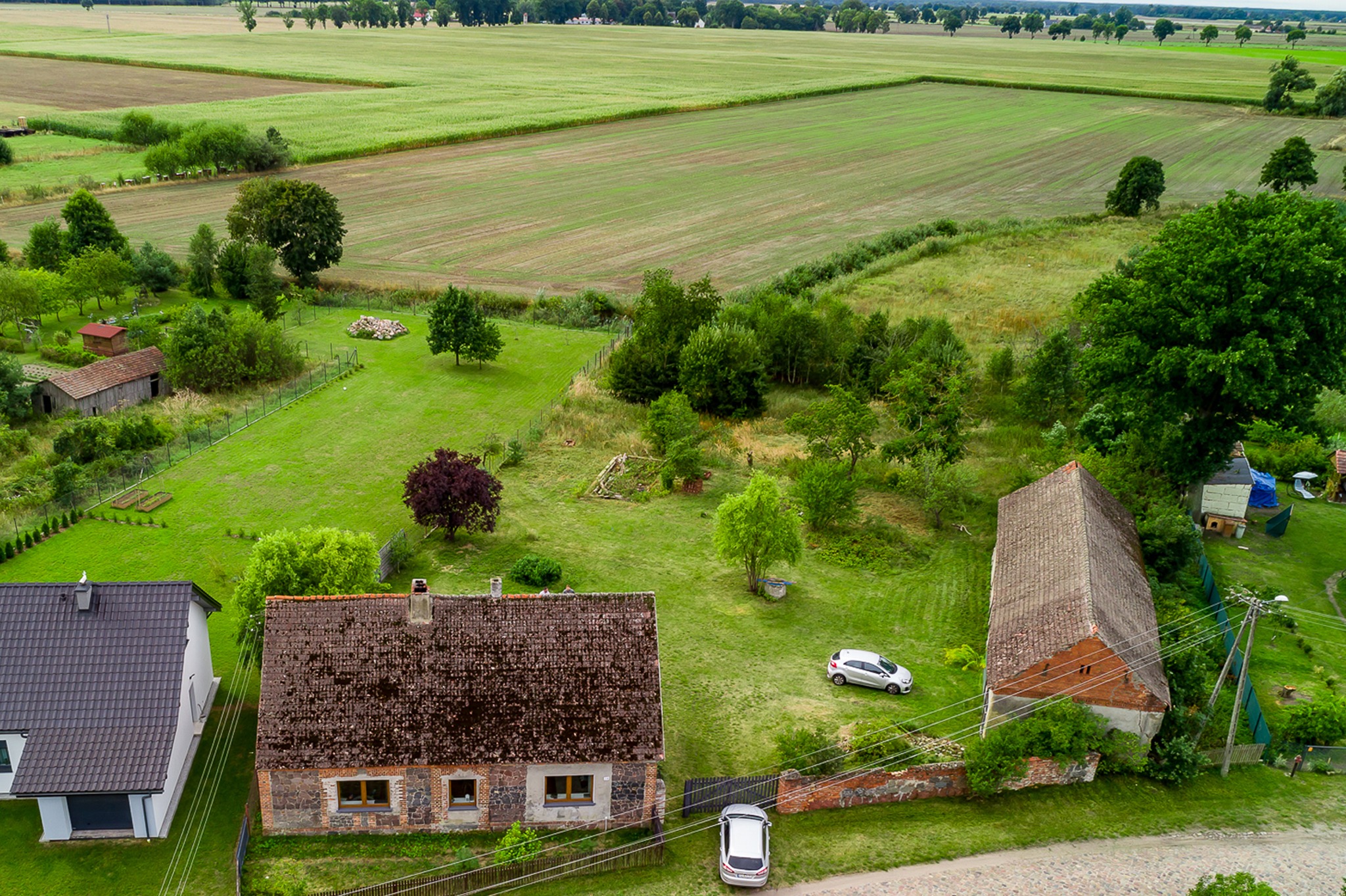 Widok z góry na dwie nieruchomości: nowoczesny dom z ciemną dachówką obok starszego domu z ceglaną dachówką pokrytą mchem, oba otoczone zielonymi trawnikami i polami w tle.