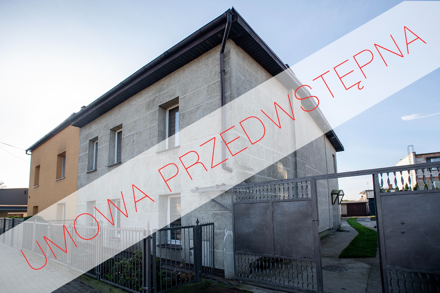 Dwupiętrowy dom z szarego betonu z ciemnym dachem, widoczny z zewnątrz przez otwartą metalową bramę, z nałożonym tekstem 'Umowa Przedwstępna'.