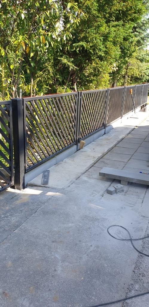 Częściowo ukończone, czarne, skośne ogrodzenie panelowe z widocznymi elementami montażowymi na betonowej podmurówce, z zielonym drzewostanem w tle.