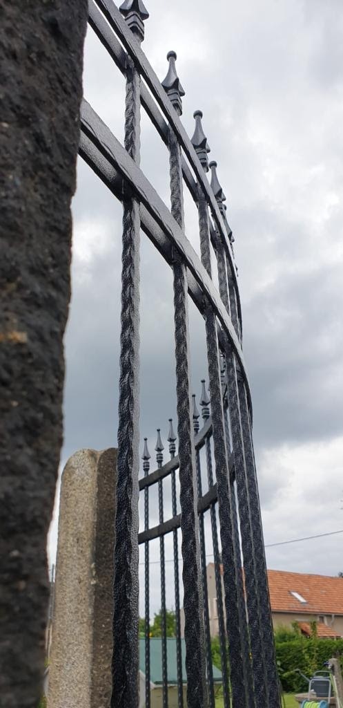 Kute, czarne ogrodzenie z dekoracyjnymi grotami, widok z dołu na pochmurne niebo, z fragmentem kamiennego słupka po lewej stronie.