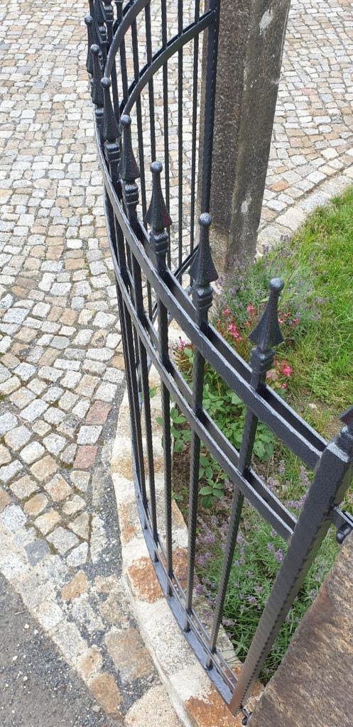 Fragment czarnego, metalowego ogrodzenia z dekoracyjnymi grotami, przylegającego do kamiennego murku, z kostką brukową i zielenią w tle.