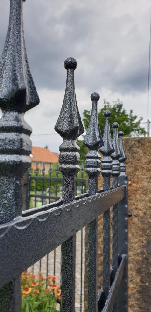 Metalowe ogrodzenie z dekoracyjnymi grotami w odcieniach szarości, widok z bliska na tle pochmurnego nieba i fragmentu kamiennego muru.