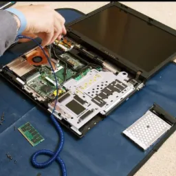 Otwarty laptop z widocznymi podzespołami, serwisant w mankiecie koszuli używa wkrętaka, niebieski kabel uziemiający podłączony do płyty głównej, obok leży dysk twardy i pamięć RAM.