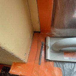 PATRYK CZEMNY - Przygotowanie narożnika ściany do prac wykończeniowych: pomarańczowa taśma uszczelniająca, paca COTERA i folia ochronna na podłodze. Detal z bliska.