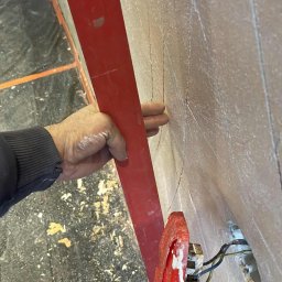 PATRYK CZEMNY - Ręka przytrzymuje czerwoną łatę na szpachlowanej ścianie z zaznaczonymi liniami, obok widoczne gniazdko elektryczne. Podłoga zabezpieczona folią.