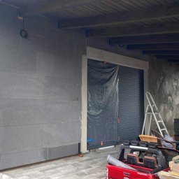 PATRYK CZEMNY - Ocieplanie ściany zewnętrznej budynku z widoczną warstwą izolacyjną i fragmentem elewacji po nałożeniu tynku. Częściowo zasłonięta brama garażowa.