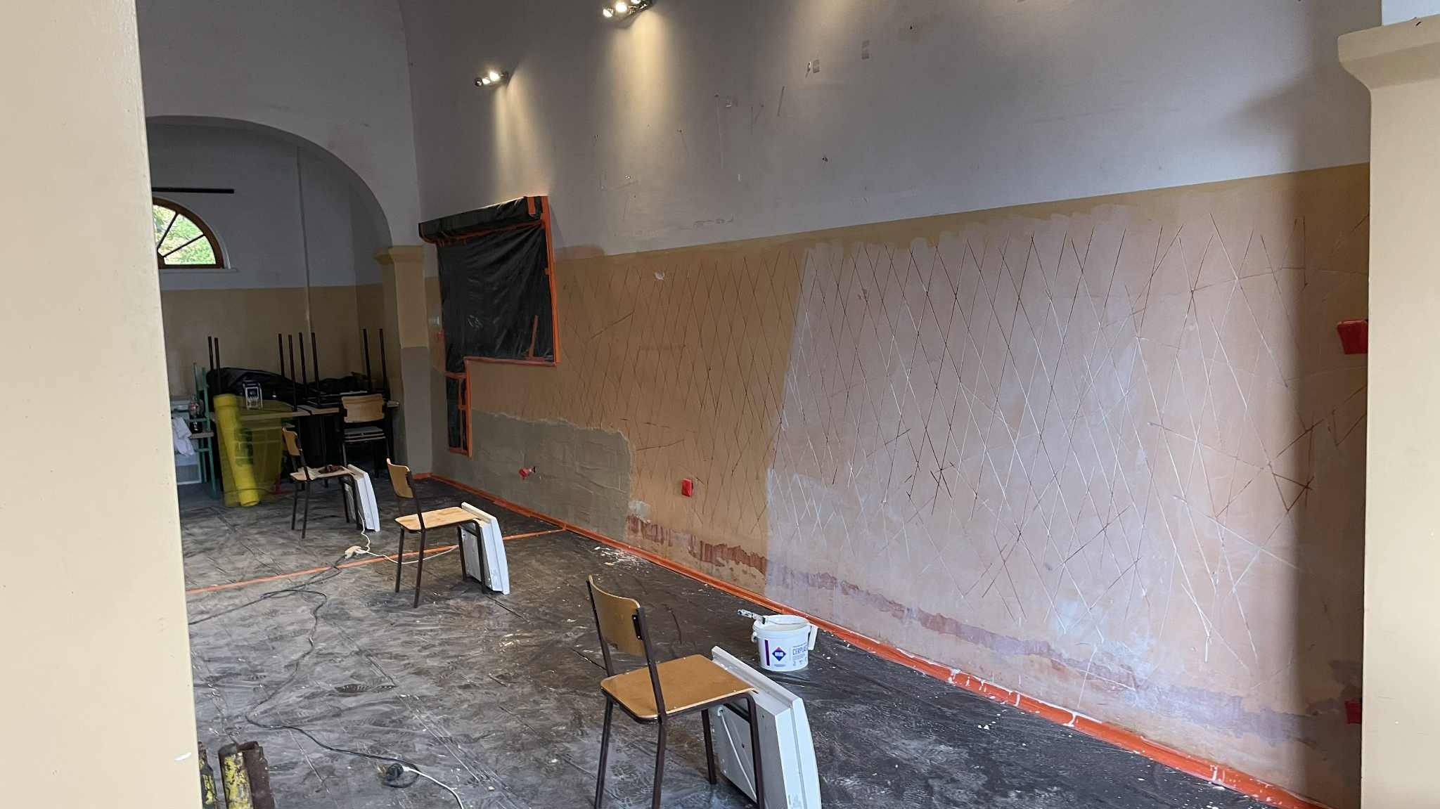 Wnętrze w trakcie remontu: ściana z nałożoną siatką i częściowo zagruntowaną powierzchnią, przygotowana do dalszych prac wykończeniowych. Widoczne krzesła i narzędzia.