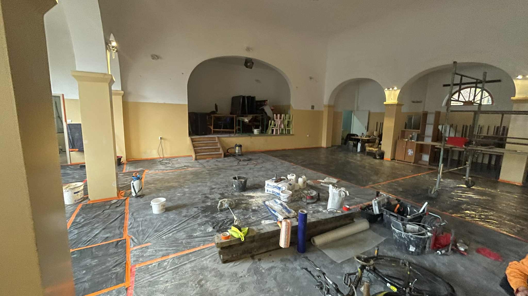 Wnętrze sali w trakcie remontu: podłoga zabezpieczona folią, widoczne narzędzia, materiały budowlane, rusztowanie i scena w tle. Przygotowania do prac wykończeniowych.