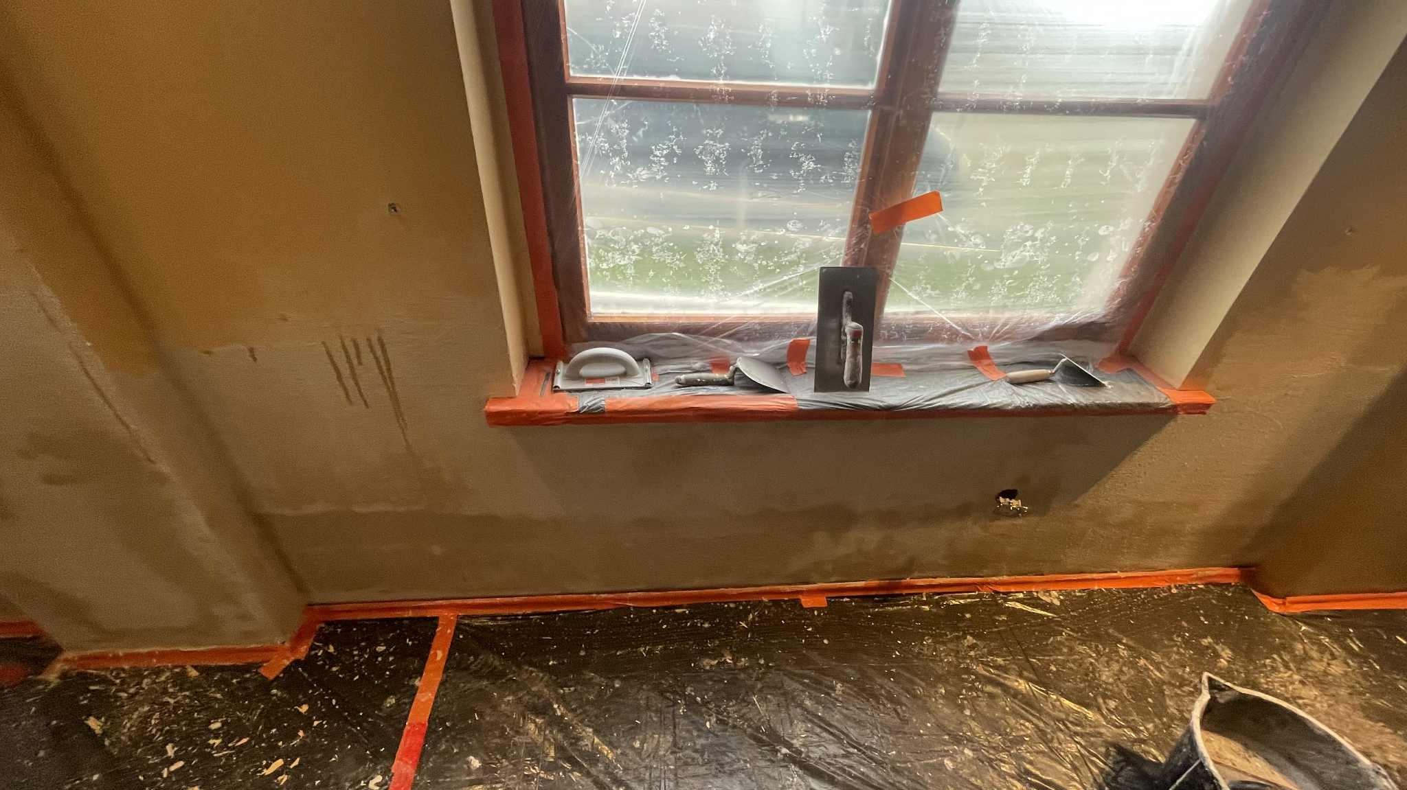 Ściana po nałożeniu gładzi szpachlowej, widoczne narzędzia na parapecie okna zabezpieczonego folią. Podłoga również zabezpieczona folią malarską.