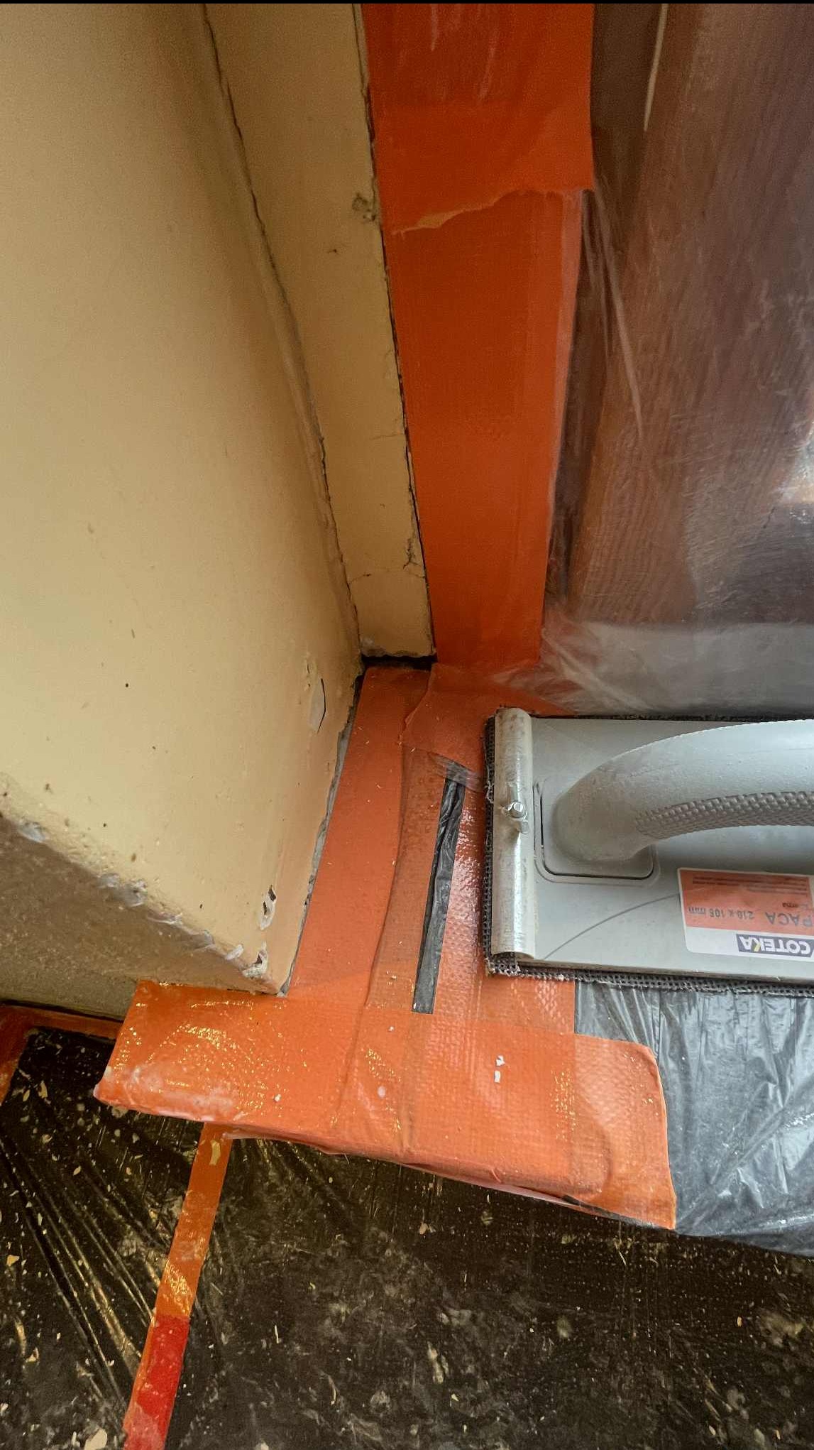 Przygotowanie narożnika ściany do prac wykończeniowych: pomarańczowa taśma uszczelniająca, paca COTERA i folia ochronna na podłodze. Detal z bliska.