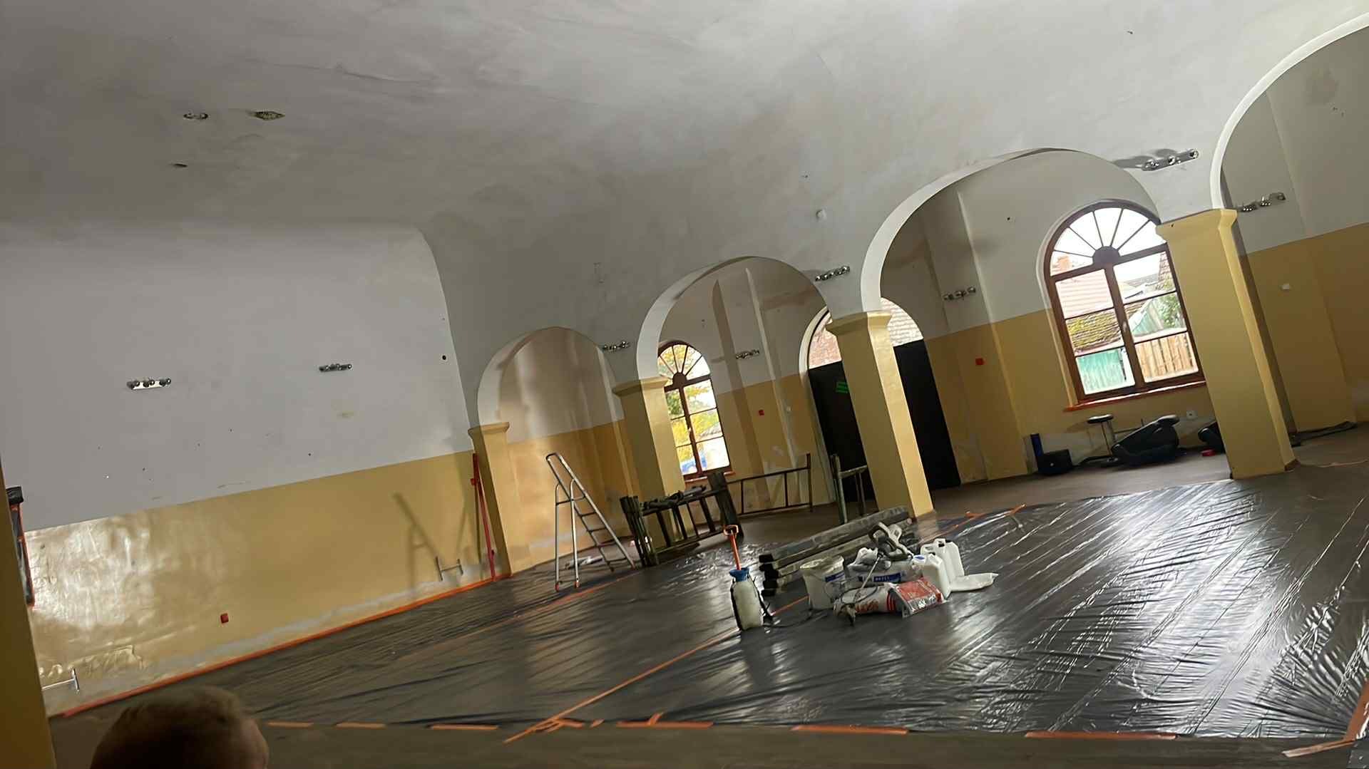 Przygotowanie do remontu dużej sali z łukami: zabezpieczona folią podłoga, drabina, narzędzia malarskie. Widoczne okna i kolumny. Etap prac remontowych.