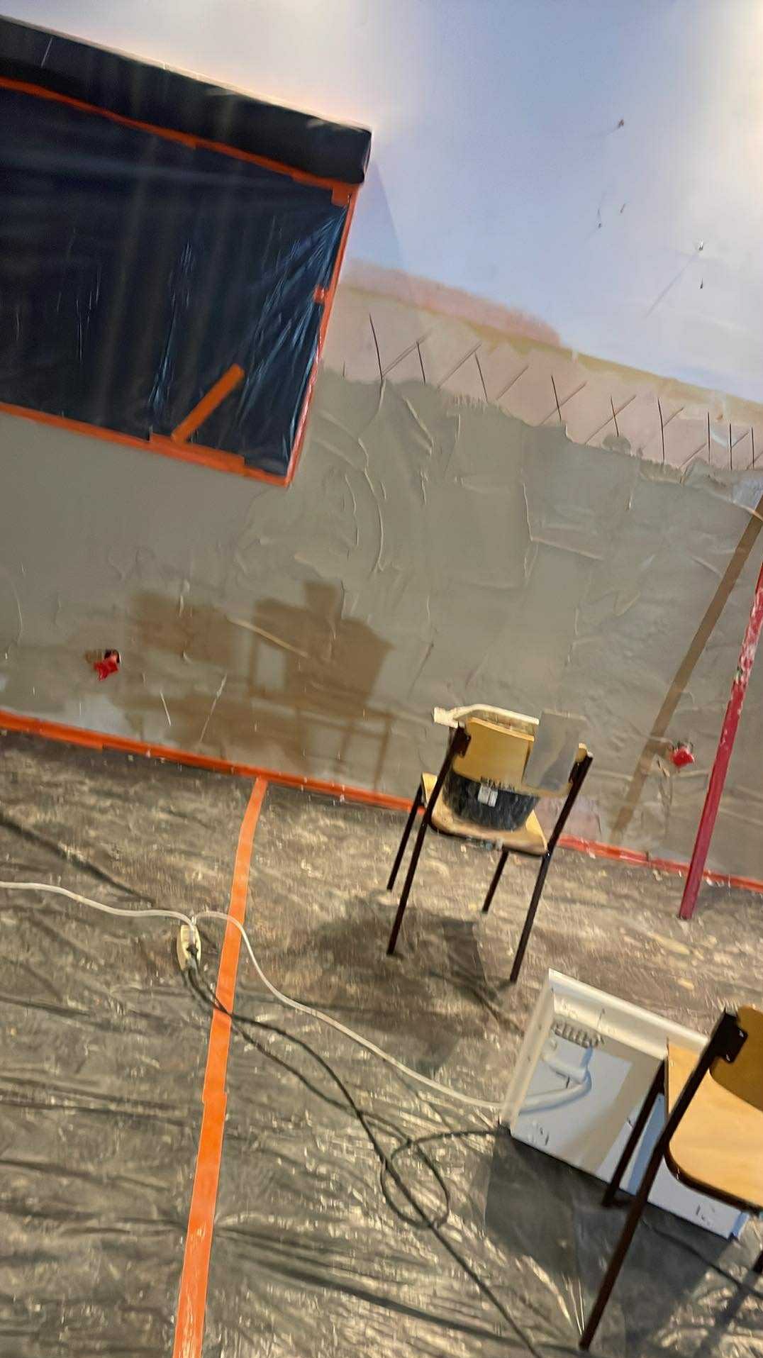 Ściana w trakcie szpachlowania, zabezpieczona folią i taśmą malarską. Widoczne narzędzia i krzesła. Prace wykończeniowe wewnątrz pomieszczenia.