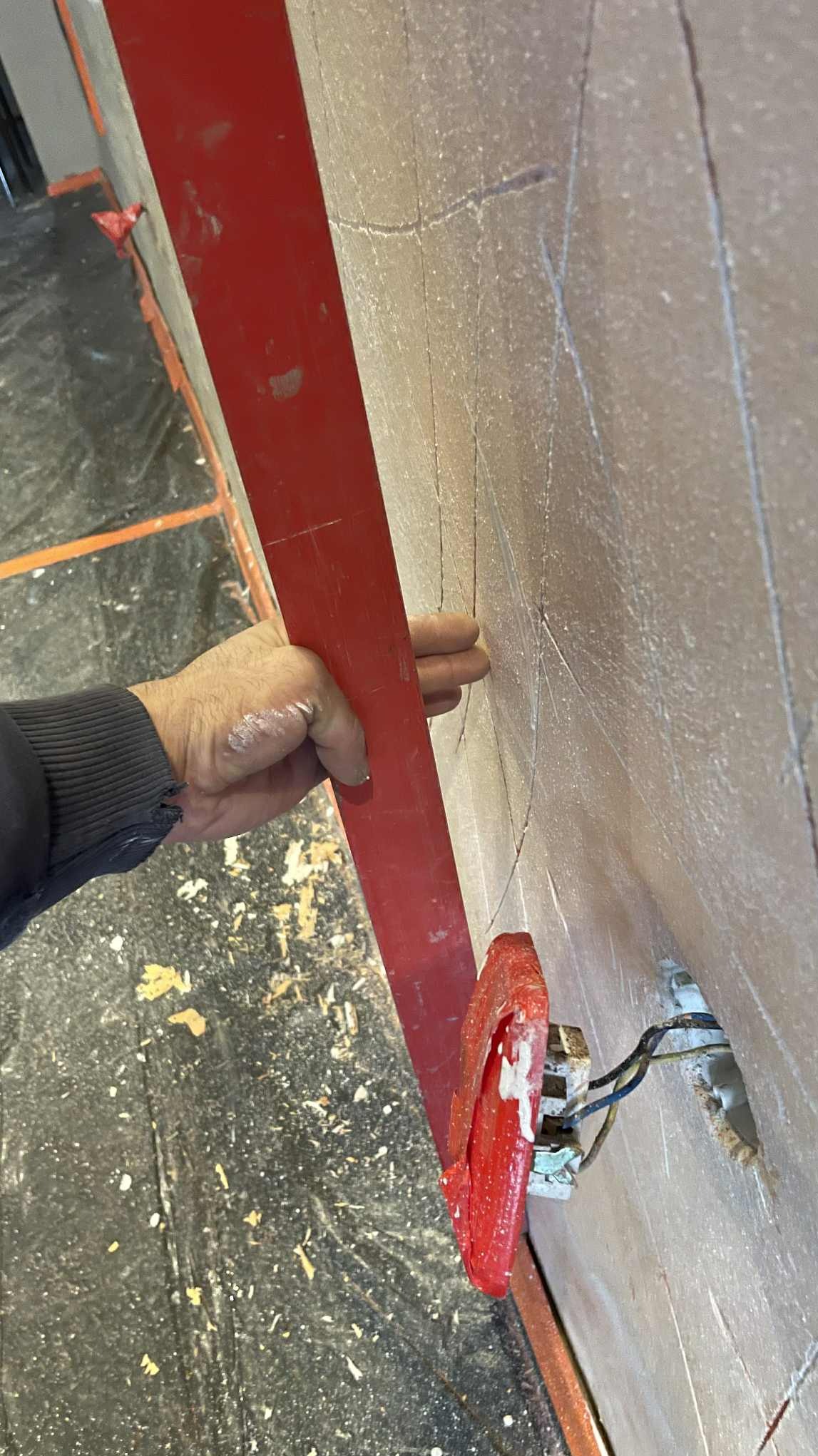 Ręka przytrzymuje czerwoną łatę na szpachlowanej ścianie z zaznaczonymi liniami, obok widoczne gniazdko elektryczne. Podłoga zabezpieczona folią.