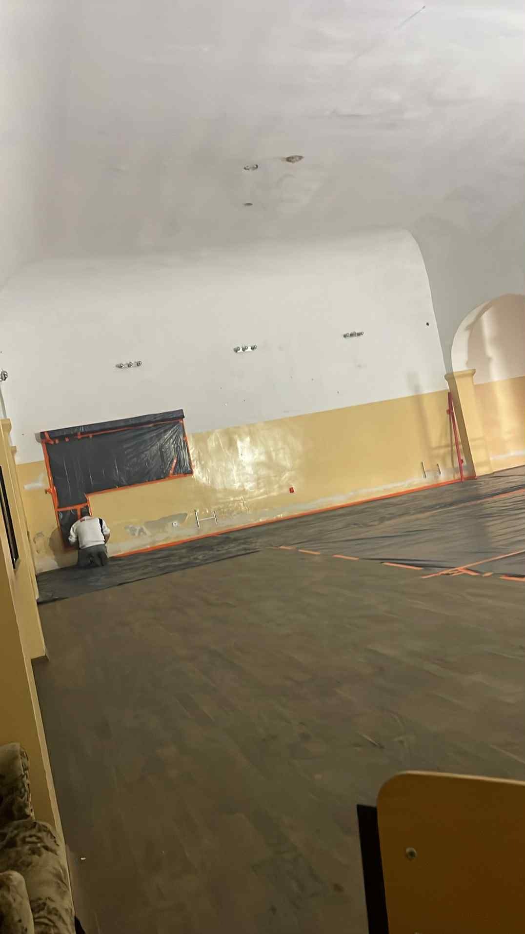 Prace przygotowawcze w dużej sali: zabezpieczanie okna folią i podłogi. Widoczny pracownik w trakcie wykonywania zadania. Pomieszczenie z łukowym sufitem.
