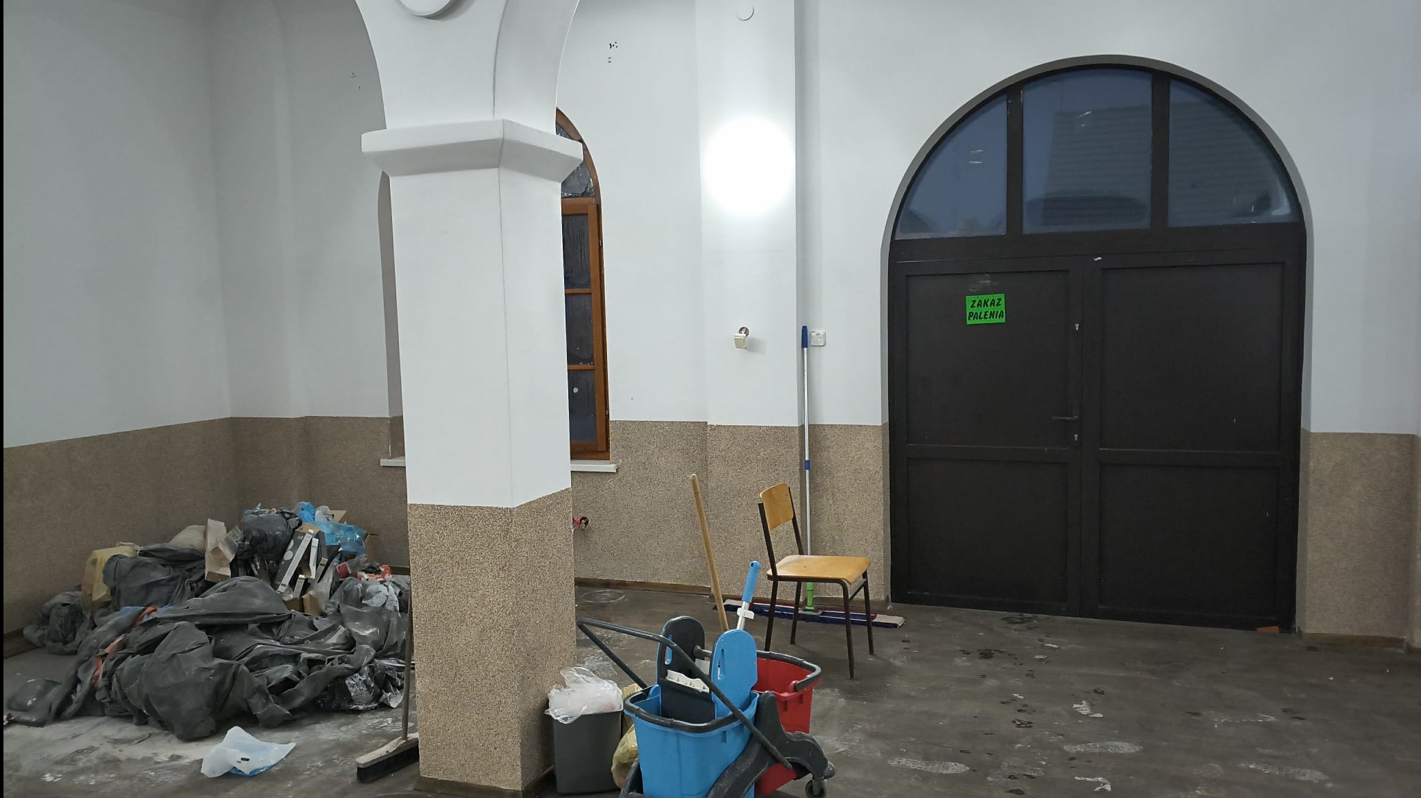 Wnętrze w trakcie remontu: worki z gruzem, wiadro z mopem, krzesło. Widoczne fragmenty kolumn i łuków. Na drzwiach tabliczka 'Zakaz Palenia'.