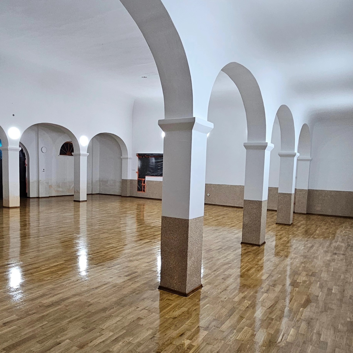 Odnowiona sala z parkietem i kolumnami. Świeżo polakierowana podłoga odbija światło. Widoczne łuki architektoniczne i fragment okna. Część ściany w trakcie prac wykończeniowych.