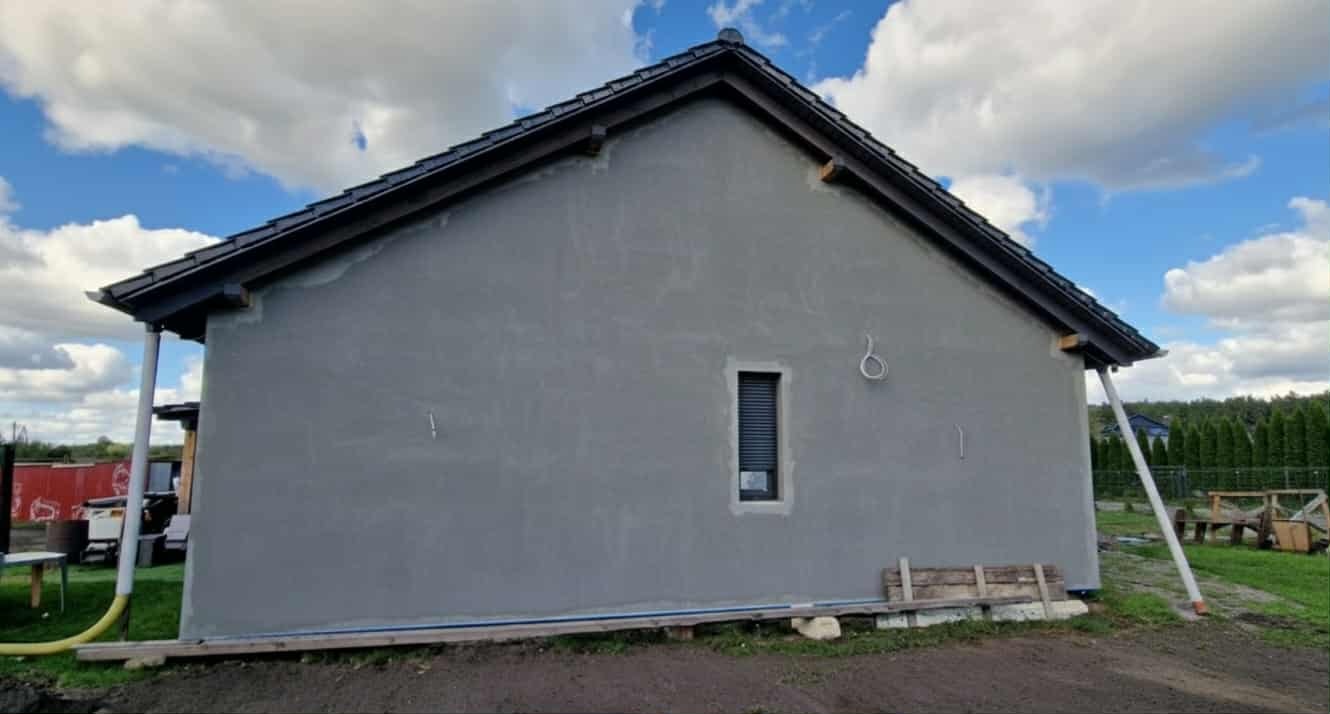 Ściana domu po nałożeniu szarego tynku, widoczne okno z roletą, fragment dachu z ciemną dachówką i podpory. Tło: błękitne niebo z chmurami.